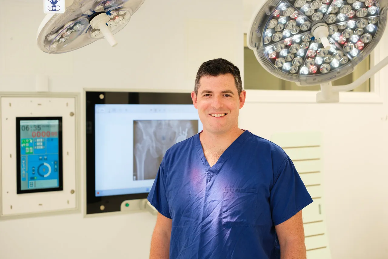 Mr James Berwin in theatre in blue scrubs, a hip X-ray on the screen behind him.